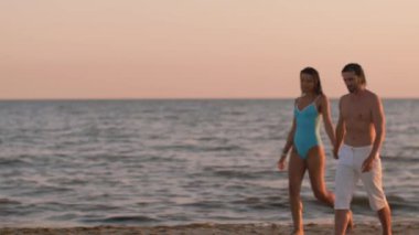 Romantik bir çift yürüyor ve gün batımında kumlu sahilde sarılıp öpüşüyor ve arkada okyanus var. Sinema yavaş çekim 4k video, reklam için tasarım ve sosyal medya, vlog
