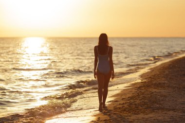 Gün batımında sahilde yürüyen siluetli bir kadın, mavi mayo giyiyor, huzurlu bir akşamın tadını çıkarıyor. Reklam için yüksek kaliteli plaj titreşimleri fotoğraf tasarımı, ve sosyal medya, vlog