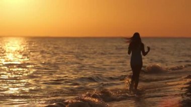 Gün batımında plajda mavi mayo giymiş, neşeyle koşan siluetli bir kadın. Mayolu bir manken gün batımında sevinçle sahile atlıyor. Kaygısız ve mutlu bir anı kutluyor.