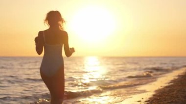 Gün batımında sahilde koşan siluetli bir kadın, mavi mayo giymiş, huzurlu bir akşamın tadını çıkarıyor. Sinema yavaş çekim 4k video, reklam için tasarım ve sosyal medya, vlog