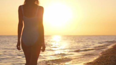 Gün batımında sahilde yürüyen siluetli bir kadın, mavi mayo giyiyor, huzurlu bir akşamın tadını çıkarıyor. Sinema yavaş çekim 4k video, reklam için tasarım ve sosyal medya, vlog