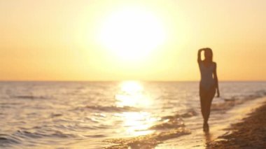 Gün batımında sahilde yürüyen siluetli bir kadın, mavi mayo giyiyor, huzurlu bir akşamın tadını çıkarıyor. Sinema yavaş çekim 4k video, reklam için tasarım ve sosyal medya, vlog