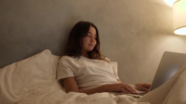 Genç bir kadın rahat bir şekilde yatakta uzanıyor, geceleri yumuşak, sıcak bir ışık altında dizüstü bilgisayar kullanarak rahat bir atmosfer yaratıyor. Sinema yavaş çekim 4k video, reklam için tasarım ve sosyal medya, vlog