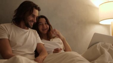 Mutlu çift yataklarında el ele tutuşup sohbet ediyorlar. Yanında da dizüstü bilgisayarları var. Sıcak ışıklandırmalı sıcak bir gece ortamı. Sinematik 4k görüntüleri, reklam için tasarım ve sosyal medya, vlog