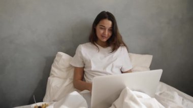 Dizüstü bilgisayarındaki bir video görüşmesi sırasında yatakta el sallayan genç bir kadın. Sabah kahvaltısı yakınlardayken rahat ve rahat bir sabah. Sinema yavaş çekim 4k video, reklam için tasarım ve sosyal medya, vlog