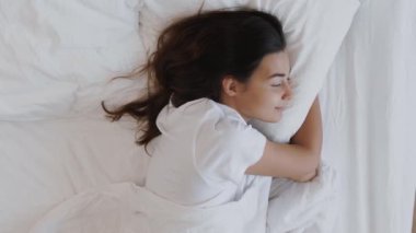 Bir kadın beyaz bir yatakta uyurken huzurlu bir şekilde gülümser, huzurlu ve rahat bir dinlenme anı yakalar, sonra uyanır ve iş için uyuyakaldığını fark eder.