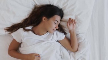 Bir kadın yatakta uyanırken gülümsüyor ve güne huzurlu ve mutlu bir başlangıç yapıyor. Uzun siyah saçlı genç kadın beyaz çarşaflarla yatakta uzanıyor, gülümsüyor ve yan tarafa bakıyor.