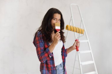 Elinde boya silindiri ve fırçayla gülümseyen genç bir kadın, DIY ev yenileme projesine hazır. Ev geliştirmesi, iç tasarım ve yaratıcı kavramlar için ideal