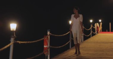 Beyaz elbiseli bir kadının aydınlatılmış bir iskelede gece vakti yürürken çekilmiş yavaş çekim görüntüleri. Seyahat blogları, romantik sahneler ve yaşam tarzı promosyonları için mükemmel..
