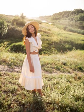 Hasır şapka takan ve açık yaz elbisesi giyen genç bir kadın manzaralı kırsal bir çayırda sıcak güneş ışığının tadını çıkarıyor. Yaz temaları, rahatlama ve doğal güzellik için mükemmel, yaşam tarzı için ideal.