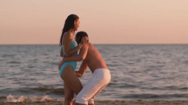 Romantik plaj aşkı. Neşeli çift gün batımında sahilde eğleniyor, beyaz pantolonlu adam mavi mayo giyen kadını kaldırıyor ve etrafında dönüyor onlar gülerken ve okyanusun kenarında akşamın tadını çıkarırken.