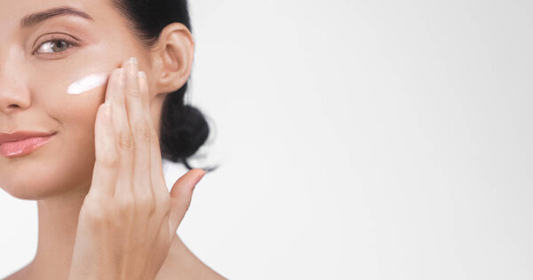 Крупный план молодой белой женщины, наносящей крем для кожи на лицо, с большим количеством места для копирования на светлом фоне. Перфект для красоты, скинхедов и велнес-рекламщиков. Высокое качество