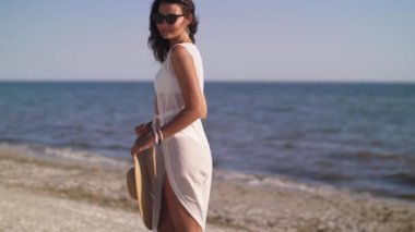 Beyaz bikinili zarif, bronzlaşmış bir kadın güneşli bir sahilde elinde güneş şapkasıyla yürüyor. Bu görüntü yaz rahatlığını ve lüksü temsil ediyor. Moda, seyahat ve yaşam tarzı reklamları için mükemmel..