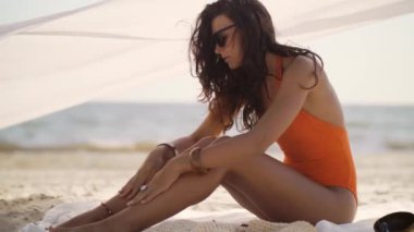 Turuncu mayo giymiş bir kadın plajda otururken güneş kremi sürerken görülüyor. Bu kaliteli video yaz, seyahat ve güzellik promosyonları için mükemmel. Güneş güvenliği ve rahatlama için.