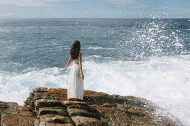 Dalgalar etrafını sararken dalgalı beyaz elbiseli bir kadın kıyı kayalıklarında okyanusa bakıyor. Görüntü denizin dinamik gücünü ve çarpıcı bir güzellikteki kadının huzurunu yakalıyor.