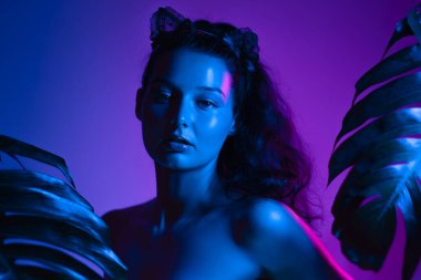 Bir kadın, çarpıcı neon mavi ve mor ışıklarla aydınlatılmış tropikal yapraklarla çevrili duruyor. Dramatik renkler modern, gizemli bir atmosfer yaratıyor, gece hayatı temaları için mükemmel.