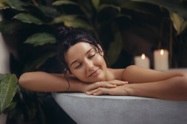 Rahat bir ifadeye sahip sakin bir kadın yemyeşil ve rahatlatıcı mum ışığıyla çevrili lüks bir banyodan zevk alır. Rahatlama, kişisel bakım ve spa reklamları için mükemmel bir kavram.