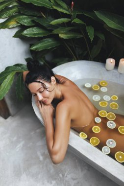 Yüzen turunçgil dilimleriyle rahatlatıcı bir banyonun keyfini çıkaran bir kadının olduğu sakin bir spa sahnesi. Yeşillik ve mumlar içeriyor. Huzurlu, yüksek kaliteli bir atmosfer yaratıyorlar.