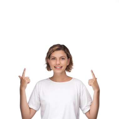 Parlak bir gülümsemesi olan neşeli genç bir kadın, iki eliyle beyaz bir arkaplanı işaret ediyor, pozitiflik, coşku ve heyecan temalarını aktarıyor. Promosyon malzemeleri için ideal
