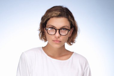 Kameraya doğrudan bakan, gözlüklü ve tişörtlü ciddi düşünceli bir kızın, gradyan mavi arka planda izole edilmiş, sorunlu bir durumu düşünen bir stüdyo fotoğrafı. Yüksek