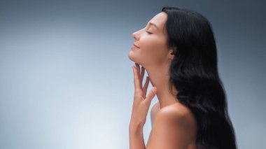 Uzun siyah saçlı zarif bir Asyalı kadın boynuna hafifçe dokunuyor, kusursuz cildini mavi bir arka plana karşı sergiliyor. Güzellik, cilt bakımı ve sağlık reklamları için ideal.