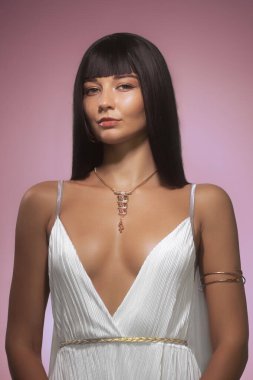 Parlak siyah saçlı, altın aksanlı beyaz bir elbise giyen ve pembe arka planda poz veren kendine güvenen bir kadın. Üst düzey moda yazıları ve lüks markalar için ideal.