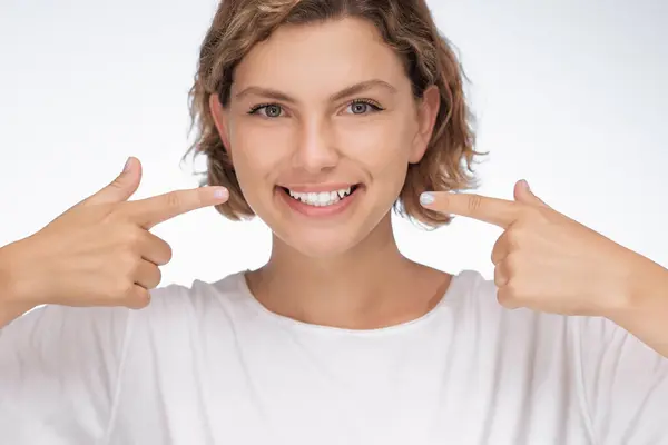 stock image A cheerful young woman with a radiant smile points at her white teeth on a white background, highlighting themes of dental health, confidence, and positivity. Perfect for dental advertisements, oral