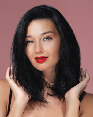 Siyah saçlı ve kırmızı dudaklı çarpıcı bir kadın pembe arka planda kendine güvenerek poz veriyor. Bu kaliteli görüntü güzellik, moda ve yaşam tarzı promosyonları için mükemmeldir.