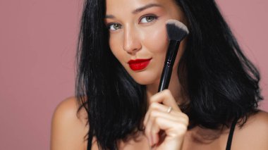 Kırmızı dudaklı ve siyah saçlı çekici bir kadın pembe arka planda duran bir fırça kullanarak makyaj yapıyor. Güzellik dersleri, makyaj sanatçısı promosyonları ve kozmetik reklamları için ideal.