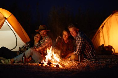 Bir grup arkadaş plaj kenarındaki kamp alanlarında kamp ateşi etrafında oturmuş turuncu çadırlarla, hikayelerini paylaşıyor ve serin bir gecede ışıltının tadını çıkarıyorlar..
