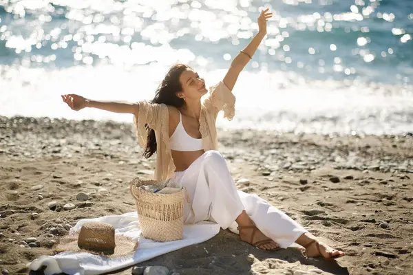 stock image A woman stretches and relaxes on a sunny beach with a straw hat and bag, enjoying the ocean breeze and serene view. Perfect for promoting travel destinations, summer fashion, and beachside leisure