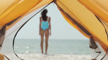 Bir çadırın içinden kumlu sahilde duran mavi mayo giymiş bir kadının arka görüntüsü. Yaz, tatil, sahil yaşam tarzı ve seyahat kavramları için mükemmel. Yüksek kaliteli yavaş çekim görüntüleri