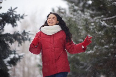 Kırmızı ceketli ve beyaz atkılı bir kadın, neşeli bir şekilde karlı bir ormanda yürüyor. Gülümsüyor ve kış gününün tadını çıkarıyor..
