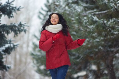 Kırmızı ceketli ve beyaz eşarplı gülümseyen bir kadın, yeşil ağaçlarla çevrili bir parkta karlı bir kış yürüyüşünün tadını çıkarıyor. Kış neşesi, açık hava aktiviteleri ve mevsimsel güzellik temaları için mükemmel.. 