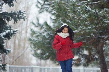 Kırmızı ceketli ve beyaz eşarplı bir kadın, yeşil ağaçlarla çevrili bir parkta karlı bir kış yürüyüşünün tadını çıkarıyor. Kış tatili, açık hava aktiviteleri ve mevsimlik eğlence için mükemmel temalar.. 