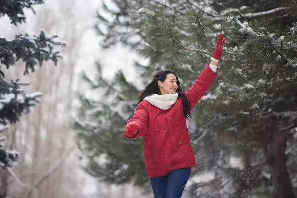 Kırmızı ceketli ve beyaz eşarplı bir kadın neşeyle Evergreen 'lerle çevrili karlı bir parkı keşfediyor. Kış mutluluğu, açık hava keşfi ve mevsimsel güzellik temaları için ideal.. 