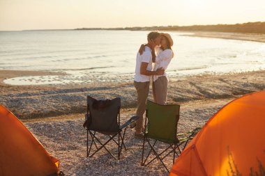 Sahildeki kamp alanlarında gün batımında turuncu çadırlarla romantik bir öpüşme yaşayan bir çift deniz kenarında mükemmel ve samimi bir an yaratıyor..