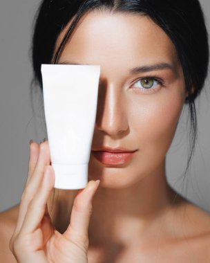 Güzel bir portrede beyaz bir cilt tüpü tutan zarif genç bir kadın. Cilt bakımı, kozmetik ve güzellik ürünü reklamları için idealdir..