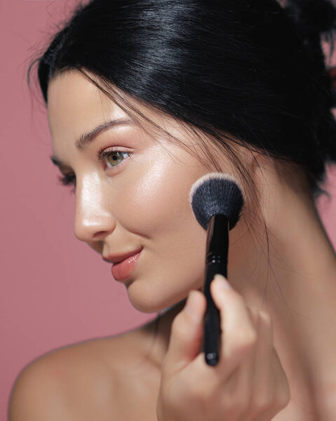 Элегантная азиатка с натуральным макияжем наносит румяна на розовый фон. Идеально подходит для продвижения красоты, скинкара и косметики, выделяя высококачественные концепции красоты. 