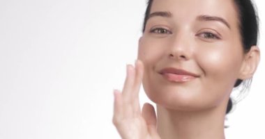 Beyaz bir arka plana hafifçe gülümseyen ve yüzüne dokunan beyaz tenli genç bir beyaz kadının yakın çekimi. Güzellik, cilt bakımı, sağlık, estetik ameliyat ve cilt beyazlatma için mükemmel.