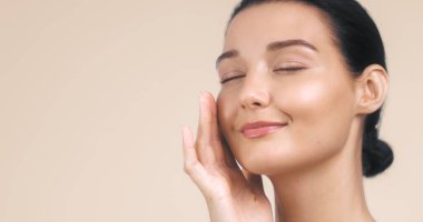 Gözleri kapalı, nazikçe yüzüne dokunan, pürüzsüz ve parlak cildini gösteren sakin bir kadının yakın çekimi. Onun barışçıl ifadesi ve yumuşak, nötr arkaplanı bu görüntüyü ideal kılıyor.