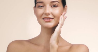 Yüzüne hafifçe dokunan, ışıl ışıl cildi olan genç ve güzel bir kadın. Tarafsız arka plan ve sakin ifadesi bu görüntüyü cilt bakımı, güzellik ve...