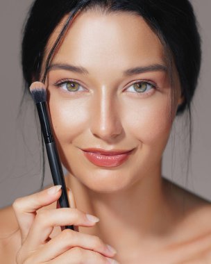 Elinde makyaj fırçası olan gülümseyen bir kadının portresi doğal güzelliğini ve parlayan cildini ön plana çıkarıyor. Güzellik, cilt bakımı, makyaj konsepti promosyonları ve yüksek kaliteli reklamlar için mükemmel..