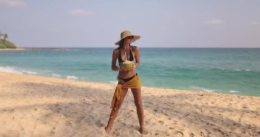 Bikini ve güneş şapkalı şık bir kadın güneşli bir tropikal plajda hindistan cevizi içeceğini seviyor. Sahne yaz gezisi promosyonları, sahil yaşam tarzı içeriği ve tatil reklamları için mükemmel..