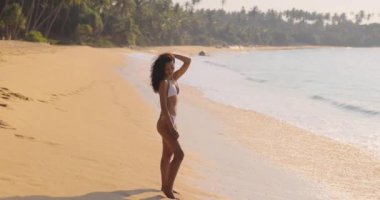 Bronzlaşmış bir kadın tropik bir plajda beyaz bir bikiniyle altın saatin sıcacık ışığında güneşleniyor. Palmiye ağaçları ve nazik dalgalarla çevrelenmiş bu görüntü lüksü, rahatlığı ve...