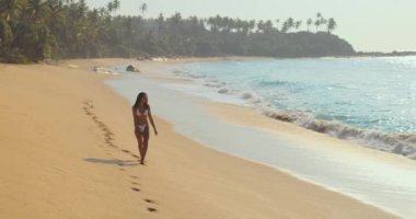 Bikinili bir kadın tenha bir tropikal sahil boyunca yürüyor ve okyanus dalgaları yavaşça kıyıya vururken kumda ayak izleri bırakıyor. Seyahat promosyonları, plaj yaşam tarzı içeriği ve huzur için mükemmel.