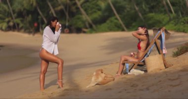 İki kadın tropik bir plajda dinleniyor, biri fotoğraf çekerken diğeri de bir köpekle sandalyede dinleniyor. Sahne kaygısız bir tatil anını yakalar, seyahat ve boş zaman kavramları için mükemmeldir..