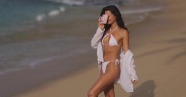 Beyaz elbiseli bir kadın güneşli bir günde tropik bir plajda kamerayla fotoğraf çekmekten zevk alır. Sahne, tatil, fotoğrafçılık ve yaz tatili ile ilgili kavramlar için ideal, boş zaman ve seyahatleri somutlaştırıyor..