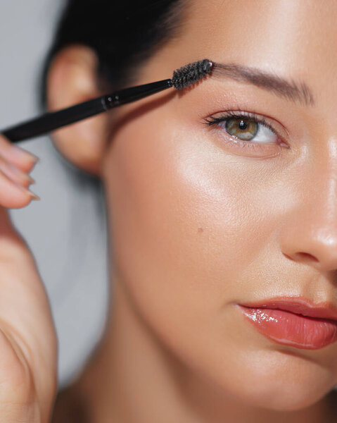 Close-up portrait of an elegant Asian woman with natural glow grooming her eyebrows. Ideal for beauty, skincare, and cosmetics advertisements, focusing on high-quality beauty concepts. 