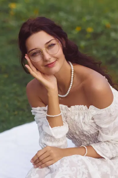 stock image A close-up portrait of a smiling woman in a white off-shoulder dress, relaxing outdoors on a blanket. The serene and elegant setting emphasizes themes of beauty, femininity, and relaxation in a